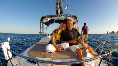 Smiling couple relaxing on a sailboat foredeck at sunset, crew member at the bow, deep blue ocean and distant rocky sea stacks — coastal sailing vacation.