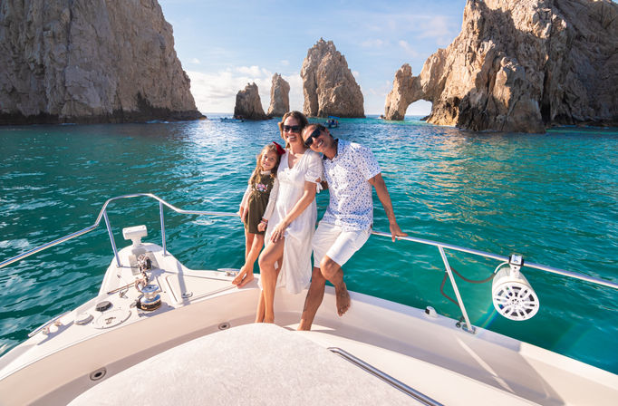 Smiling family of three on the bow of a white boat in turquoise water near dramatic coastal rock arches at Cabo San Lucas under a bright blue sky