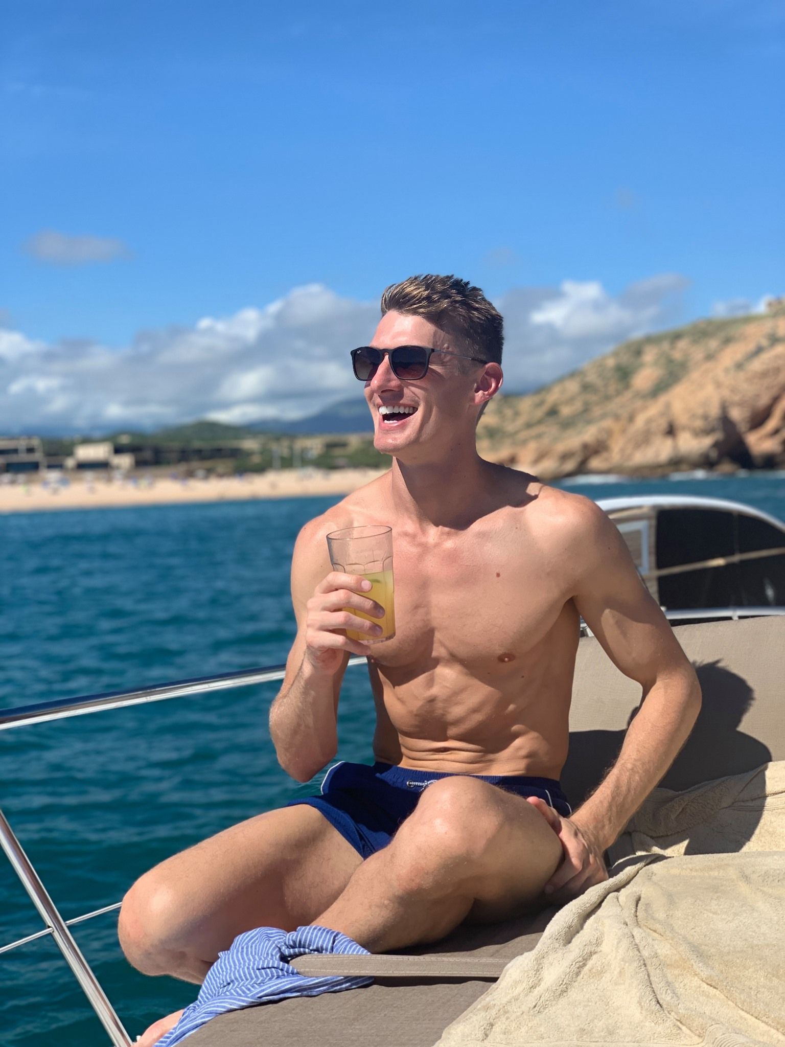 Smiling shirtless man in sunglasses relaxing on a sunlit yacht with a cold drink, turquoise ocean, sandy beach and rocky coastline under a bright blue sky — vacation vibe.
