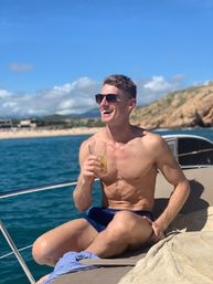 Smiling shirtless man in sunglasses relaxing on a sunlit yacht with a cold drink, turquoise ocean, sandy beach and rocky coastline under a bright blue sky — vacation vibe.