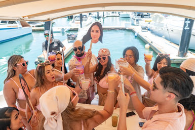 Sunlit boat party at a marina with friends in pink swimsuits and heart-shaped sunglasses raising colorful cocktails for a celebratory toast near docked yachts, playful vibe with cardboard face props.