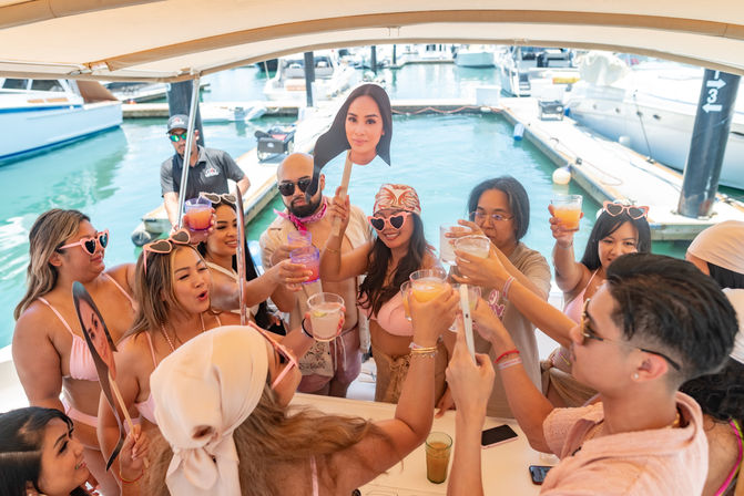 Sunlit boat party at a marina with friends in pink swimsuits and heart-shaped sunglasses raising colorful cocktails for a celebratory toast near docked yachts, playful vibe with cardboard face props.