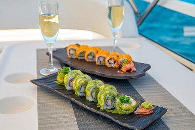 Avocado-wrapped and orange-roe-topped sushi rolls on black plates with pickled ginger and wasabi, two champagne flutes on a yacht table with blue ocean in the background