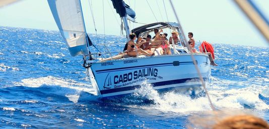 Group of people enjoying a sunlit white sailboat cutting through deep-blue Pacific waves off Cabo San Lucas, Baja California.