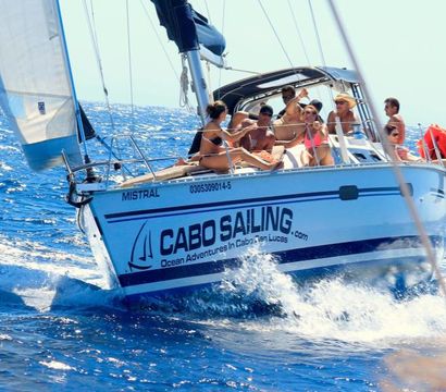 Group of people enjoying a sunlit white sailboat cutting through deep-blue Pacific waves off Cabo San Lucas, Baja California.