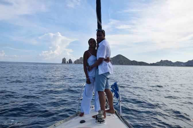 Couple hugging on the bow of a sailboat off Cabo San Lucas, Mexico, with rocky sea arches, blue ocean and a bright sky.