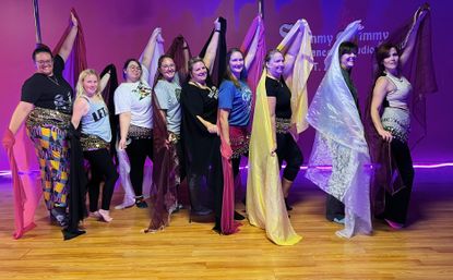 Nine dancers lined up in a purple-lit studio on a wooden floor, wearing coin hip scarves and holding colorful silk veils overhead while smiling — playful group belly dance class pose.