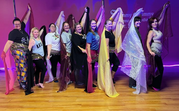 Nine dancers lined up in a purple-lit studio on a wooden floor, wearing coin hip scarves and holding colorful silk veils overhead while smiling — playful group belly dance class pose.