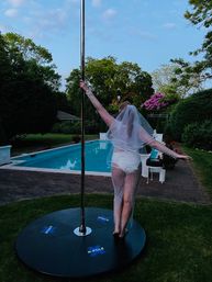 Person wearing a sheer beaded veil and shorts posing on an X-Pole in a backyard by a swimming pool at dusk, arms out amid garden trees and flowering shrubs.