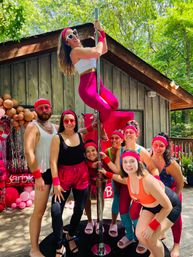 Energetic group in pink retro workout gear posing around a chrome dance pole on a sunlit wooden deck by a forested cabin, with pink balloons and party decor.