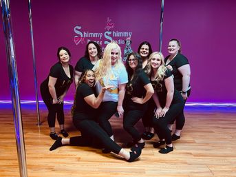 Group of women posing and smiling in a bright pink pole-fitness studio with chrome poles and hardwood floor, wearing black workout outfits around a woman with a sash — celebratory group fitness vibe.