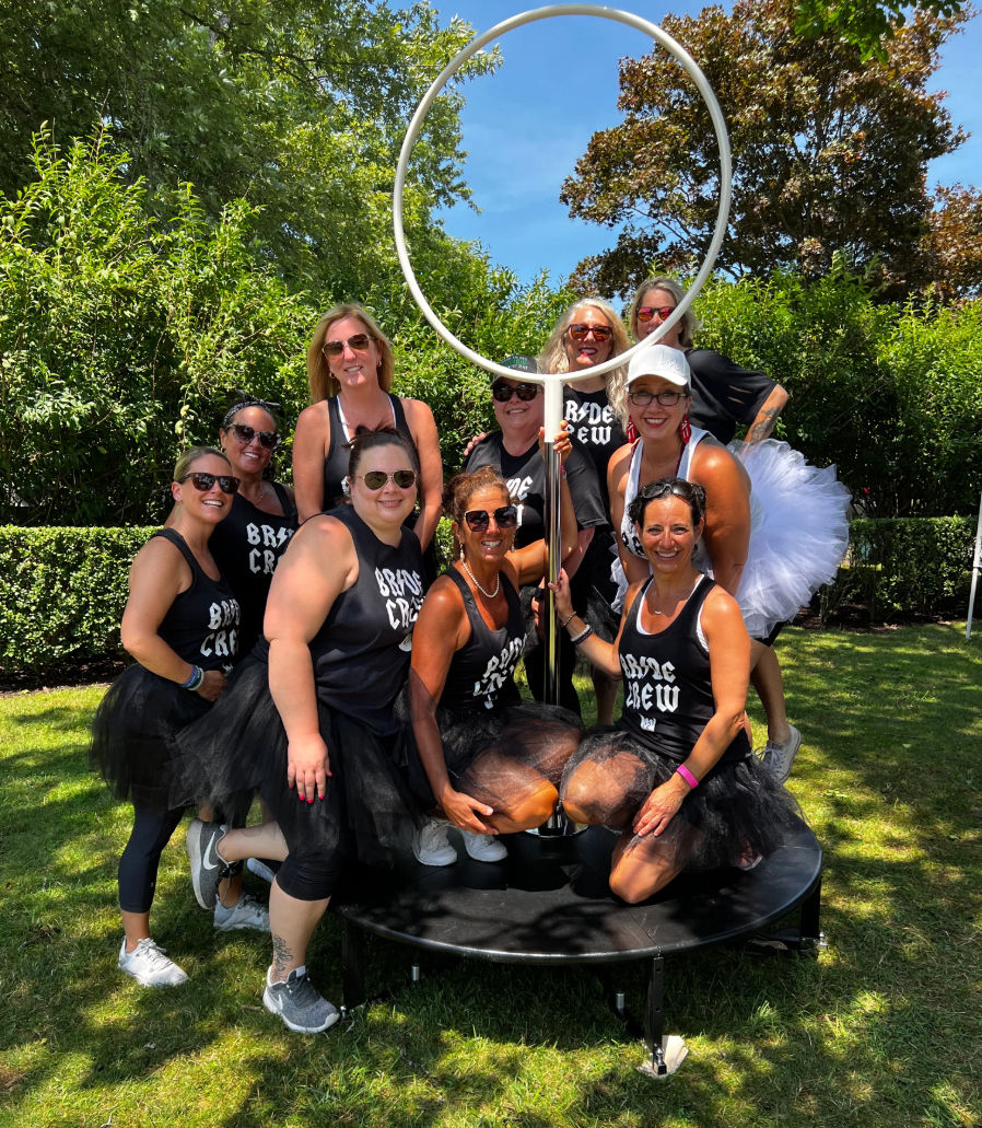 Outdoor bachelorette party group photo: smiling women in matching “Bride Crew” tanks and black tutus posing around a large circular ring prop on a sunny grassy lawn with trees.
