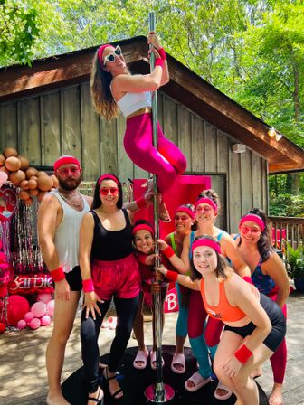 Outdoor summer cabin-deck party with friends in bright pink 80s-style workout gear posing around a dance pole — woman in white crop top and hot-pink leggings climbs the pole, pink balloons and green trees in background.