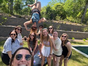 Group of friends smiling for a selfie in a sunny backyard by a pool, one woman upside-down on a pole doing a playful acrobatic pose while others laugh and pose in sunglasses with a stone terraced wall and trees behind them.