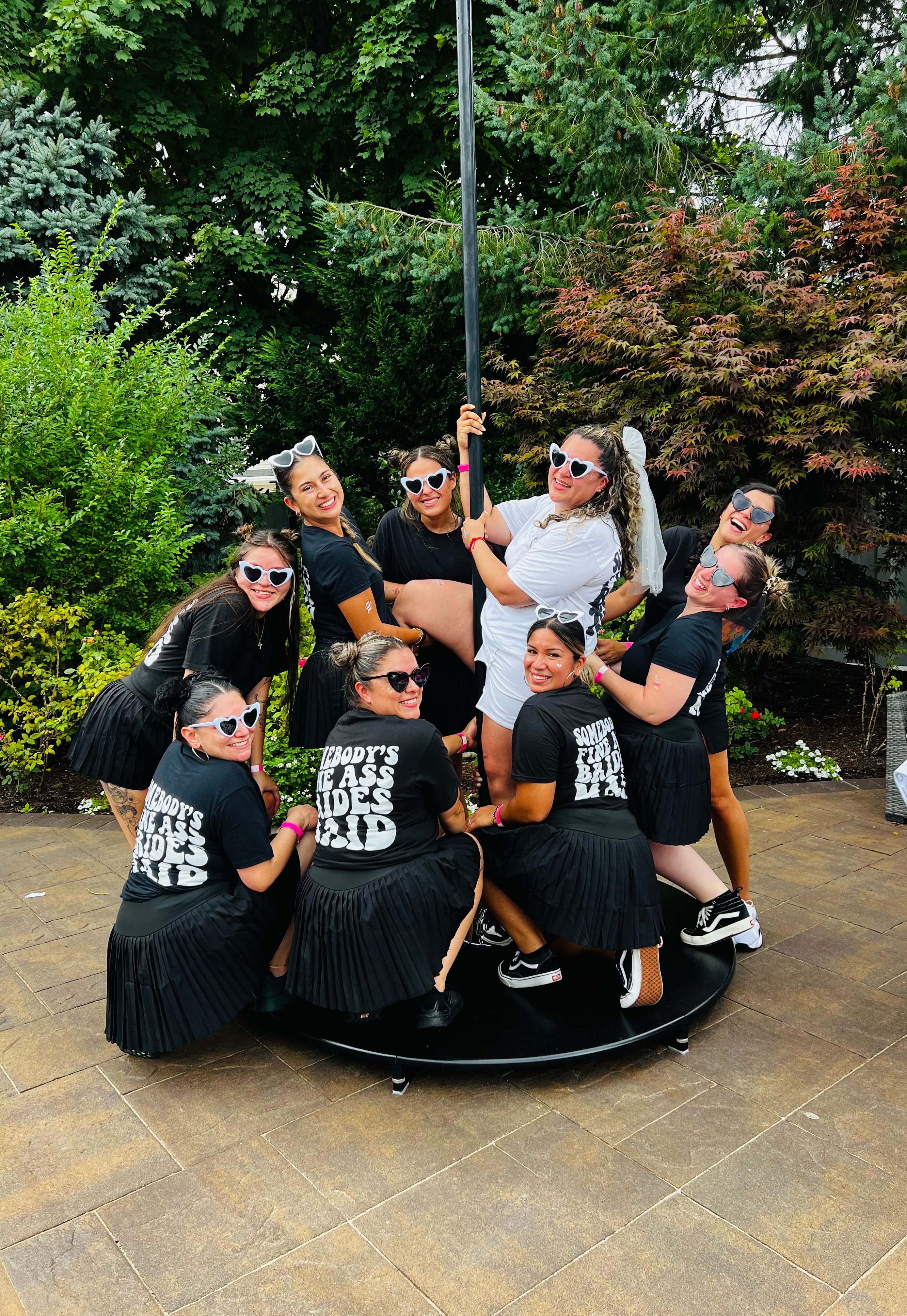Bachelorette party on a backyard patio — bride in white with a veil holding a dance pole, surrounded by friends in matching black shirts, pleated skirts and heart-shaped sunglasses, laughing among garden greenery.