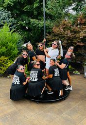 Bachelorette party on a backyard patio — bride in white with a veil holding a dance pole, surrounded by friends in matching black shirts, pleated skirts and heart-shaped sunglasses, laughing among garden greenery.