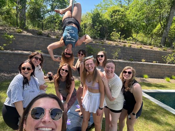 Cheerful group of women smiling at a sunny backyard pool party; one performs an upside-down pole acrobatic above the lawn, with a pool, stone retaining wall and trees in the background.