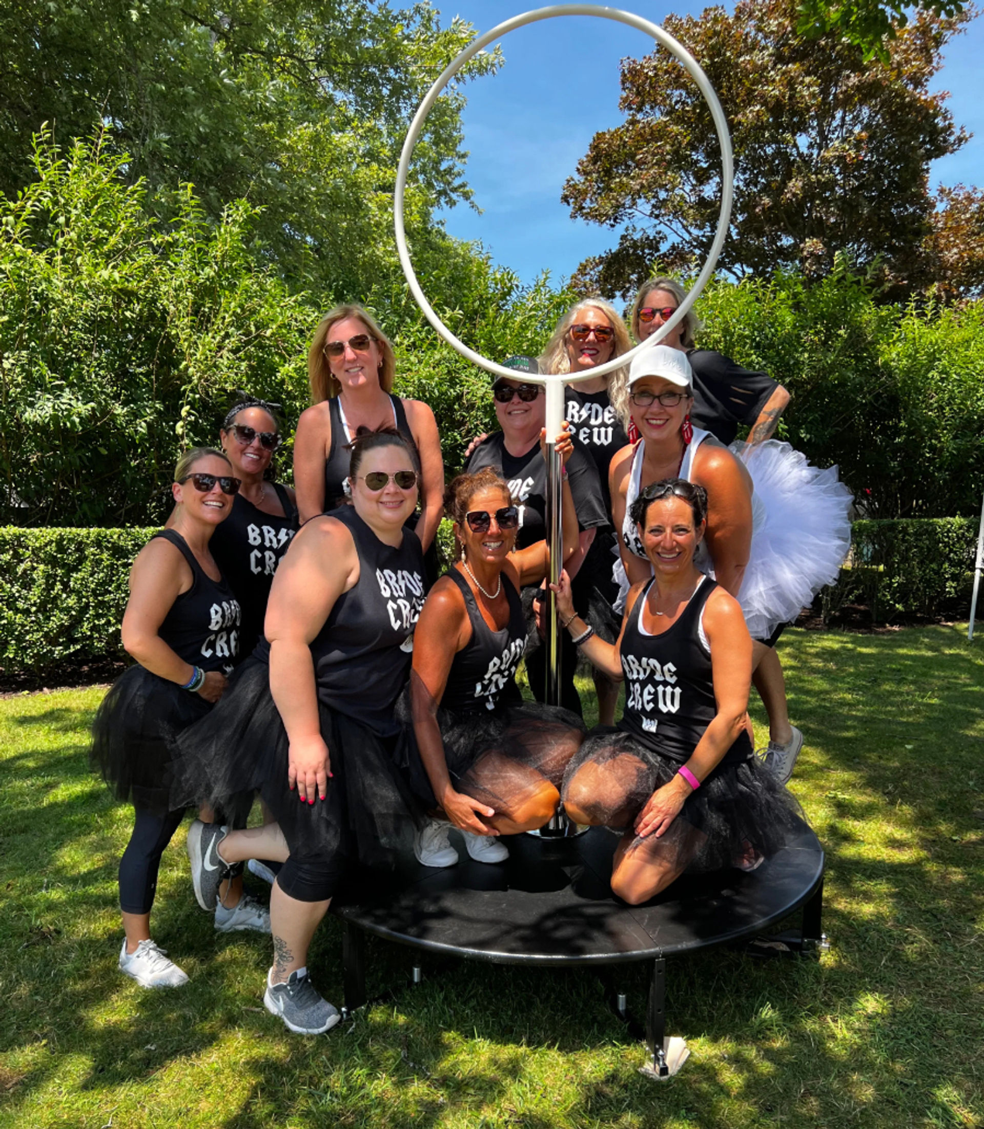 Group of women in matching Bride Crew tank tops and black tutus posing around a tall circular hoop on a small stage in a sunny park with green trees — fun outdoor bachelorette party scene
