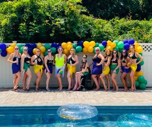Backyard summer pool party group photo of a dozen women in colorful bikinis and sarongs (purple, green, yellow) and sunglasses posing by a pool in front of a matching balloon garland with inflatable rings in the water.