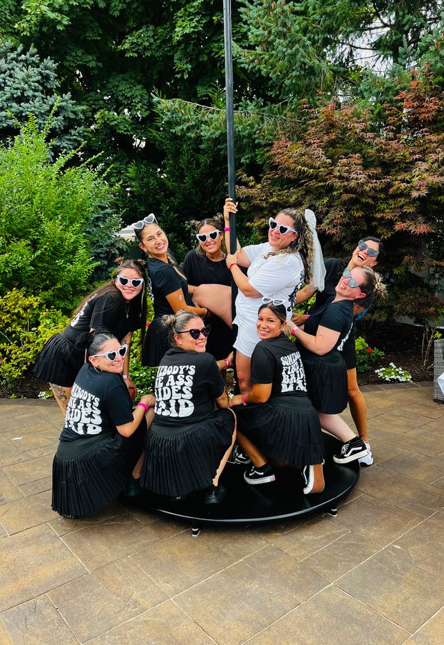 Bachelorette party on a backyard patio — women in matching black skirts and heart-shaped sunglasses pose around a pole dance stage while the bride-to-be in a white veil laughs amid lush garden greenery.