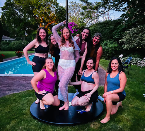 Eight women posing around a portable pole on a backyard lawn next to a swimming pool at dusk, one in a sparkly sheer outfit and others in athletic wear — outdoor pole fitness party