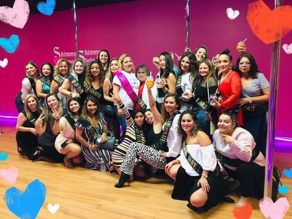 Large group of women posing and smiling in a bright pink pole-dance studio, many wearing sashes and one with a pink bridal sash holding a drink for a bachelorette party celebration.