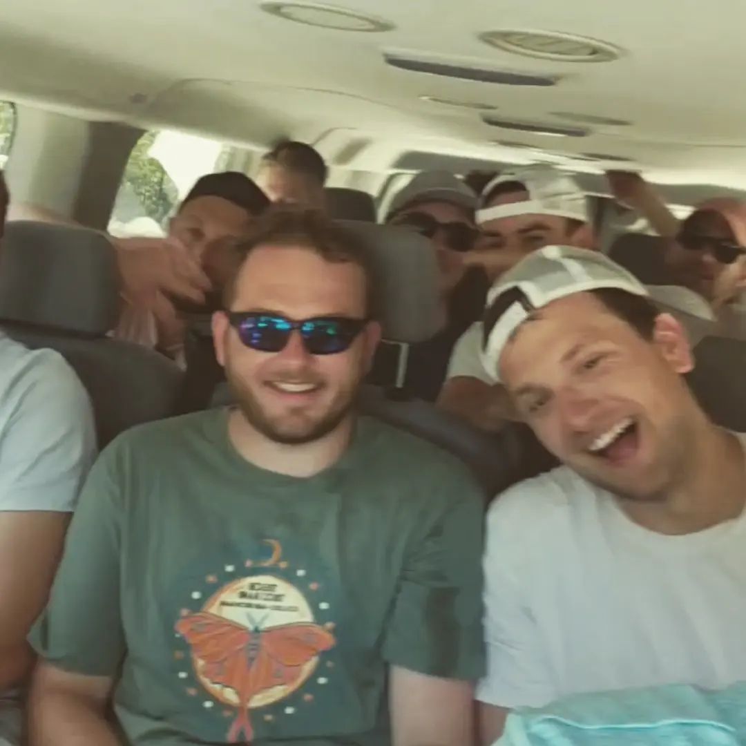 Cheerful group of young men crowded inside a passenger van on a road trip; two in front—one with blue sunglasses and a green butterfly graphic tee, the other in a backward white cap—others wearing caps and sunglasses in the back.