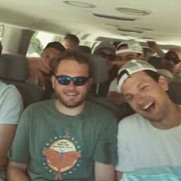 Group of cheerful young men wearing sunglasses and baseball caps packed into a passenger van, laughing during a sunny road trip.
