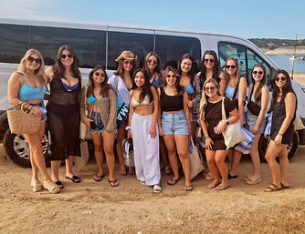 Group of friends in swimsuits and summer outfits posing by a white passenger van on a sunny shoreline with turquoise water and rolling hills