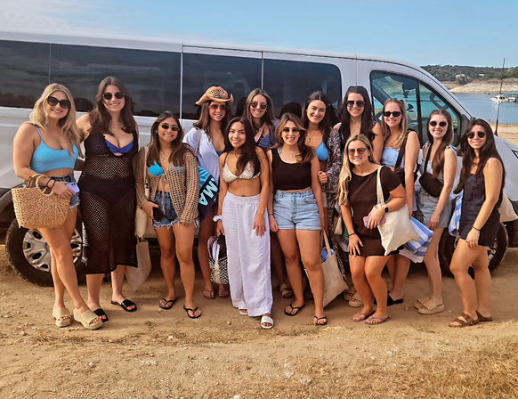 Group of friends in swimsuits and summer outfits posing by a white passenger van on a sunny shoreline with turquoise water and rolling hills