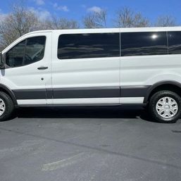 White full-size passenger van side profile parked on an asphalt lot under a blue sky with bare trees, dark tinted windows and silver wheel covers.
