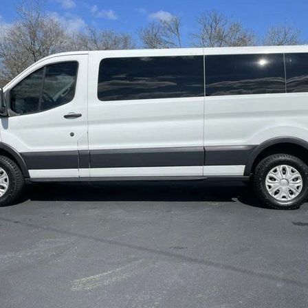 White full-size passenger van side profile parked on an asphalt lot under a blue sky with bare trees, dark tinted windows and silver wheel covers.
