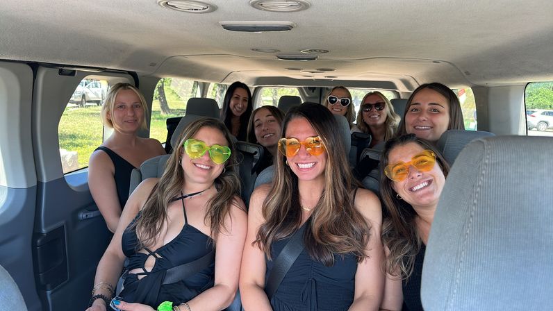 Group of women friends on a sunny road trip, packed into a passenger van and smiling—several wearing colorful heart-shaped sunglasses and casual black outfits