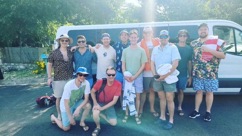 Group of friends in summer shirts, hats and sunglasses posing in front of a white passenger van on a sunny roadside, holding beach towels and backpacks for a coastal beach trip.