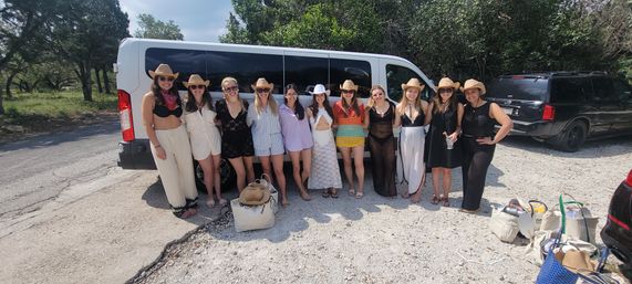 Group of women in cowboy hats and summer outfits posing in front of a white passenger van in a sunny rural parking area with trees — friends ready for a country outing.