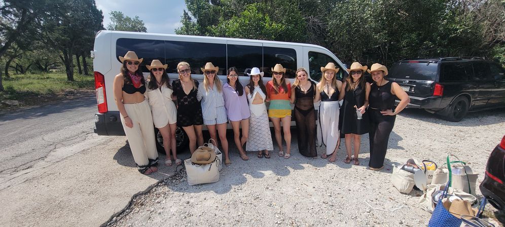 Group of women in cowboy hats and summer outfits posing in front of a white passenger van in a sunny rural parking area with trees — friends ready for a country outing.