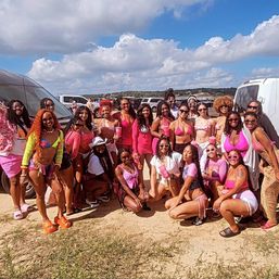 Large beach-day crew of women in pink swimsuits and summer outfits posing together on a sandy lot by parked vans under a bright blue sky with fluffy clouds.