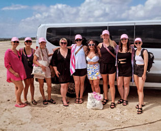 Nine women in pink caps and summer outfits posing by a white van on a sandy beach under a blue sky — sun-ready beach vacation group photo.