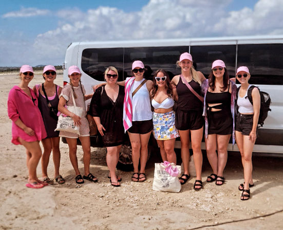 Nine women in pink caps and summer outfits posing by a white van on a sandy beach under a blue sky — sun-ready beach vacation group photo.