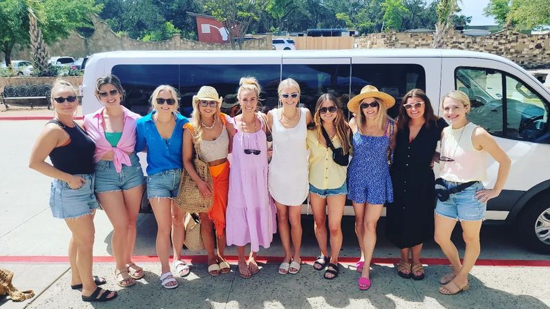 Group of women in summer outfits — shorts, sundresses, sunglasses and straw hats — smiling and posing in front of a white passenger van on a sunny sidewalk, casual vacation/girls' getaway vibe.