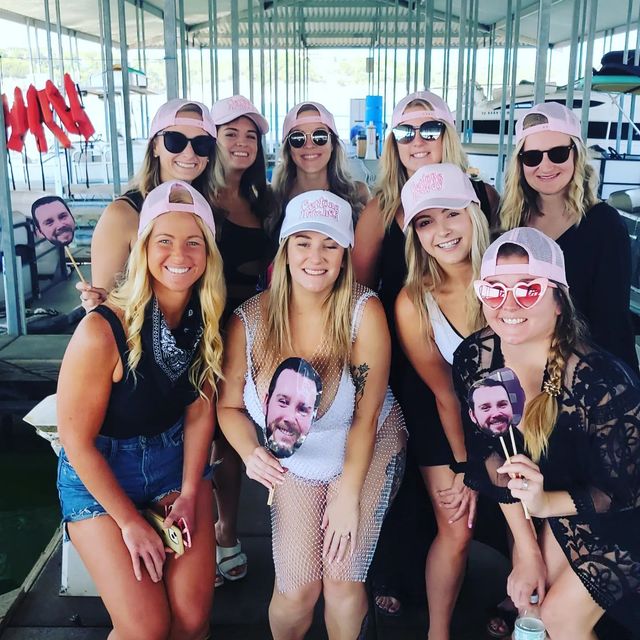 Smiling group of women in pink baseball caps and sunglasses posing on a covered lake marina dock, holding novelty face cutouts for a bachelorette party.
