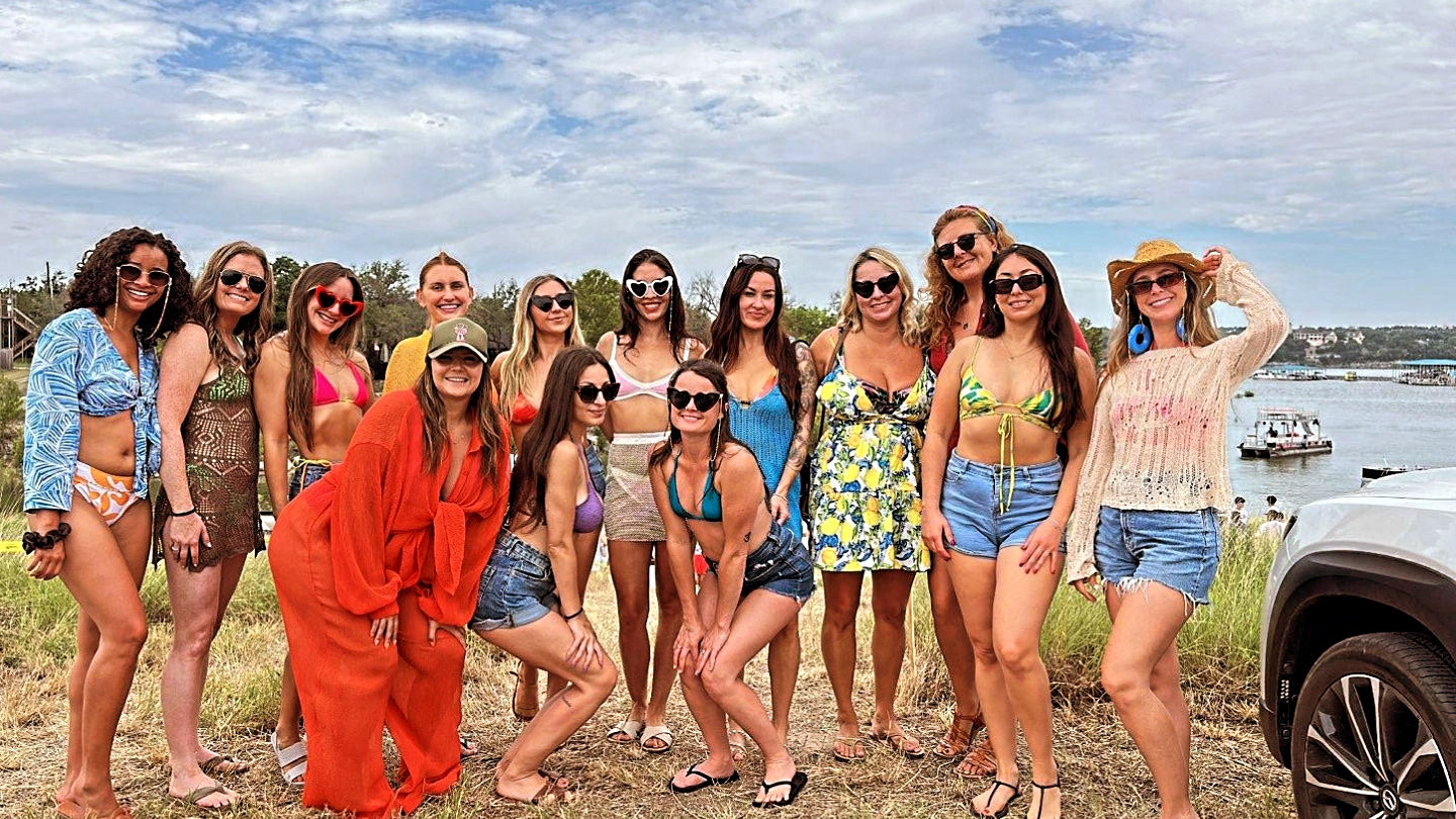 Group of women in swimsuits and summer outfits smiling and wearing sunglasses while posing on a grassy lakefront hill with boats on the water and a partly cloudy sky — outdoor summer group photo.