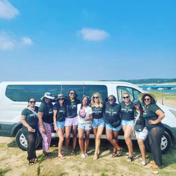 Ten women in matching black T-shirts, sunglasses and shorts posing in front of a white passenger van on a sunny lakeside beach — friends on a summer coastal van trip.