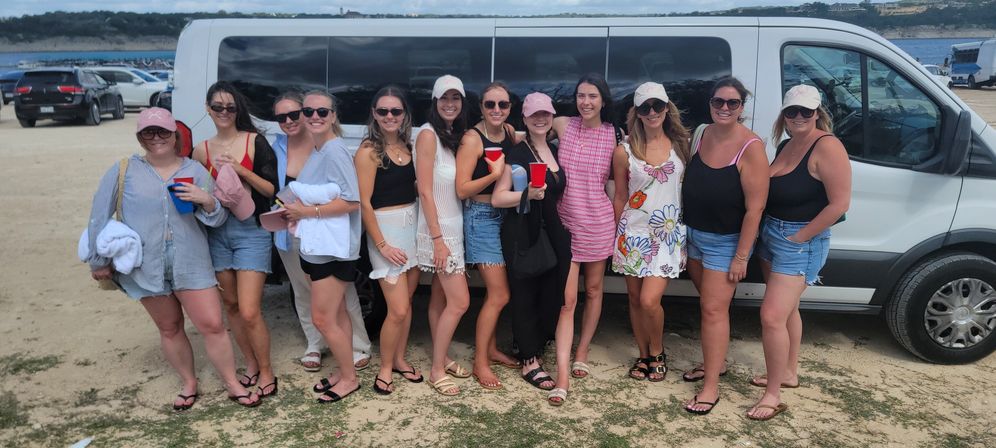 Group of smiling women in summer outfits posing in front of a white passenger van on a sandy beach parking lot with ocean and cliffs in the background, some holding red cups and towels.