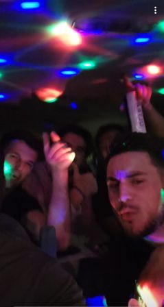 Crowded party selfie of friends indoors under multicolored disco lights, energetic nightlife vibe with one person raising a drink can.