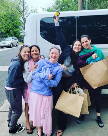 Five friends smiling and posing beside a white passenger van on a leafy suburban street, holding reusable grocery bags, water bottles and sunglasses, ready for a group outing.