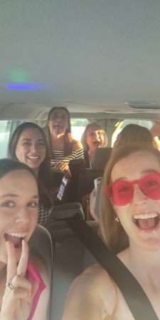 Six friends laughing and taking a selfie inside a sunlit SUV on a road trip, front passenger wearing red heart-shaped sunglasses and a seatbelt, others smiling and making playful faces.