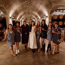 Eight women in denim and boots smiling with wine glasses under arched cellar ceilings beside oak barrels in a rustic winery wine cellar.