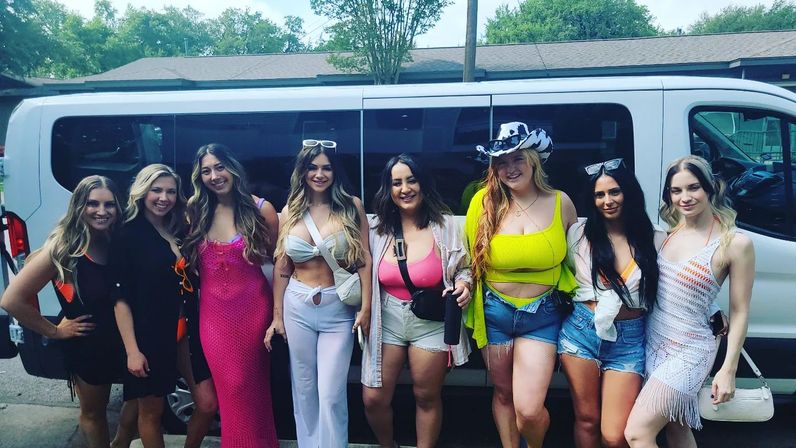 Eight women in bright summer outfits — crop tops, denim shorts, sundresses and a cowgirl hat — posing cheerfully in front of a white passenger van on a tree-lined street.