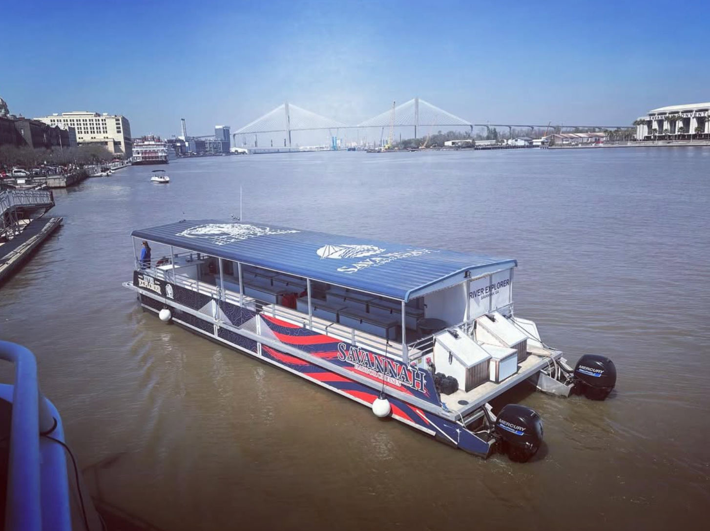 Blue-canopied riverboat on the Savannah River with a cable-stayed bridge and waterfront skyline under a clear blue sky